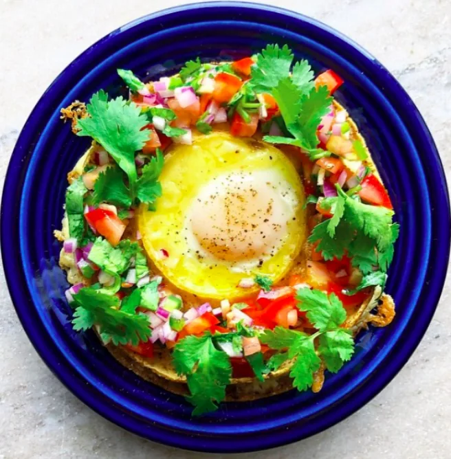 A cheesy corn tortilla stack with an egg in the center, tomato salsa and cilantro on a deep blue plate